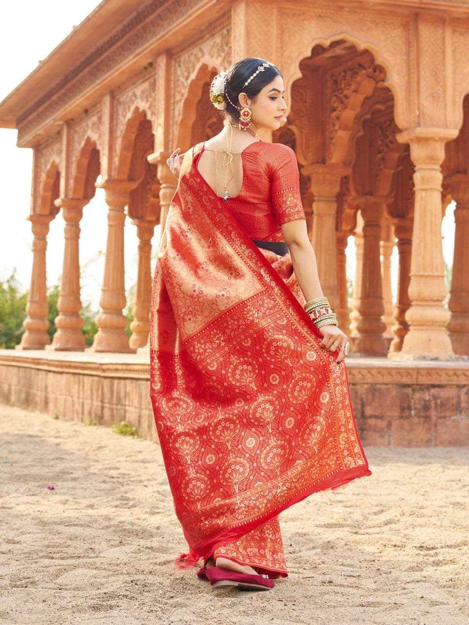 Woman in a red saree with white patterns standing in front of an architectural structure.